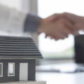 Miniatura de uma casa sobre uma mesa simbolizando um imóvel, com duas pessoas apertando as mãos ao fundo, representando a conclusão de um acordo imobiliário ou transação de compra e venda.