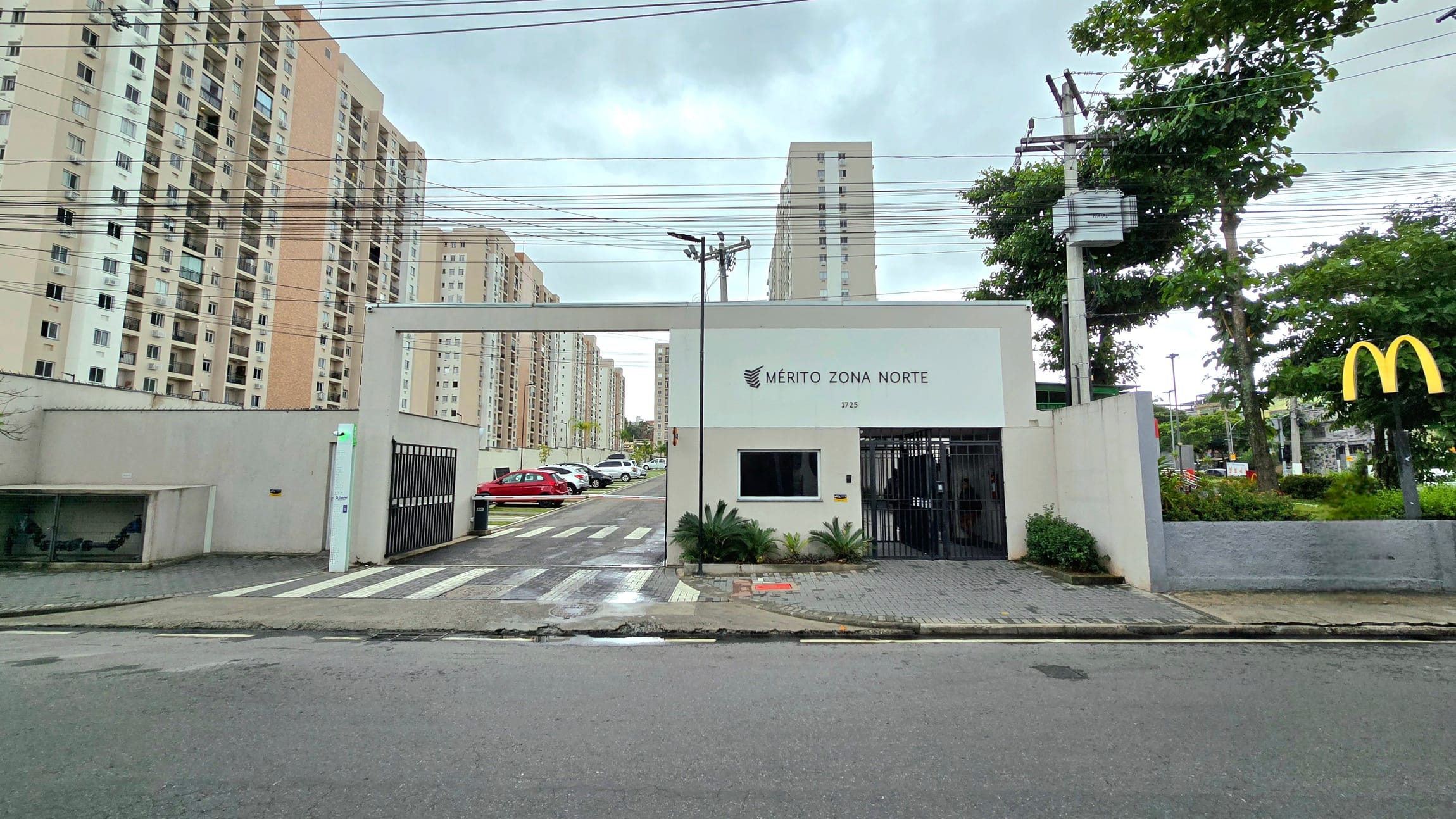 Fachada do condomínio Mérito Zona Norte, mostrando a portaria com guarita branca, o nome do condomínio e o número 1725 na parede. Ao fundo, diversos prédios altos em tons de bege e branco. À direita da imagem, há árvores e o símbolo amarelo do McDonald’s. O céu está nublado e o chão úmido, indicando que choveu recentemente.